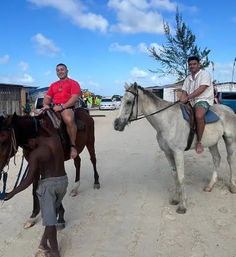 Two smiling riders on horseback trotting along a sandy coastal village street under a bright blue sky, with wooden buildings and parked cars in the background.