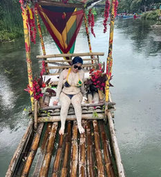 Person in bikini and sunglasses covered in mud relaxing on a decorated bamboo raft with colorful canopy and flowers, drifting on a calm tropical river