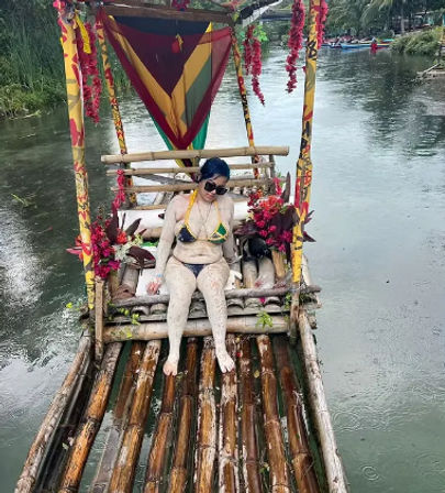 Person in bikini and sunglasses covered in mud relaxing on a decorated bamboo raft with colorful canopy and flowers, drifting on a calm tropical river