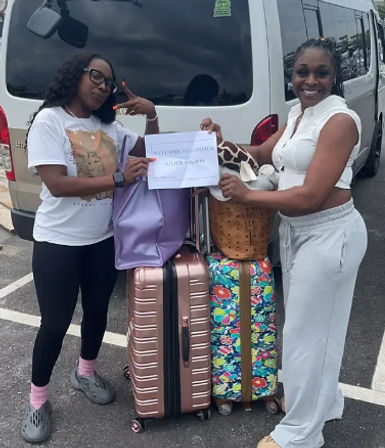 Two travelers smiling by a white shuttle van holding a "Welcome to Jamaica" paper sign above stacked colorful suitcases and bags in a parking lot.