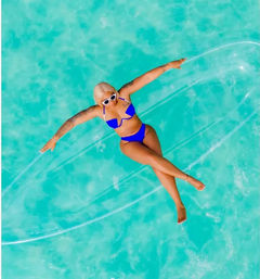 Aerial view of a woman in a cobalt blue bikini and white sunglasses lounging on a clear inflatable float in crystal turquoise pool water