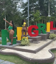 Two women in bright bikinis posing on large colorful tourist letter sign in a tropical park with palm trees