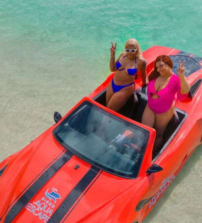 Two women in colorful swimsuits flashing peace signs while seated in a red convertible-style jet boat in shallow clear turquoise water over a sandy seabed, tropical beach vibe.