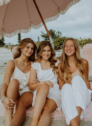 Three smiling friends lounging under a pink-and-white striped fringe beach umbrella on a sandy seaside with coastal trees and houses in the background