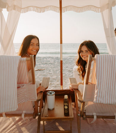 Two friends smiling in a striped beach cabana, sitting in wooden chairs with a table holding sunscreen and a tumbler, facing calm ocean waves at golden hour.