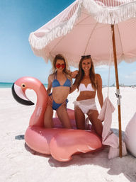 Two smiling women in bikinis posing on a giant pink flamingo float under a fringed pink-striped beach umbrella on white sand with turquoise ocean and clear blue sky.