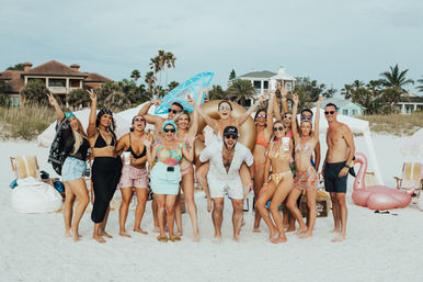 Group of friends at a beach party on white sand with palm trees and beachfront houses, wearing swimsuits, holding drinks and inflatable floats, cheering for the camera