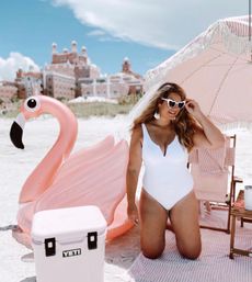 Beach vacation scene: woman in a white one-piece and heart-shaped sunglasses kneels under a pink striped umbrella beside a giant pink flamingo float and white cooler, with resort buildings in the background.