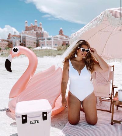 Beach vacation scene: woman in a white one-piece and heart-shaped sunglasses kneels under a pink striped umbrella beside a giant pink flamingo float and white cooler, with resort buildings in the background.