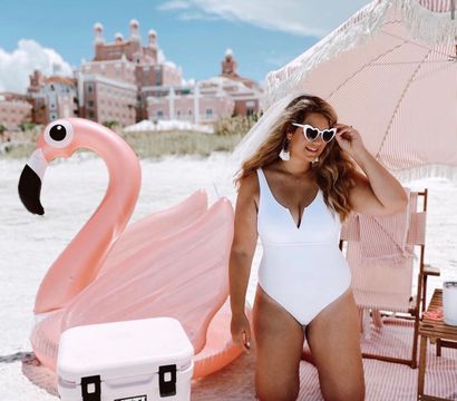 Beach vacation scene: woman in a white one-piece and heart-shaped sunglasses kneels under a pink striped umbrella beside a giant pink flamingo float and white cooler, with resort buildings in the background.