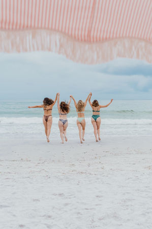 Four friends in bikinis holding hands and running toward the turquoise ocean on a white sandy beach beneath a red-and-white striped fringe umbrella under a cloudy sky
