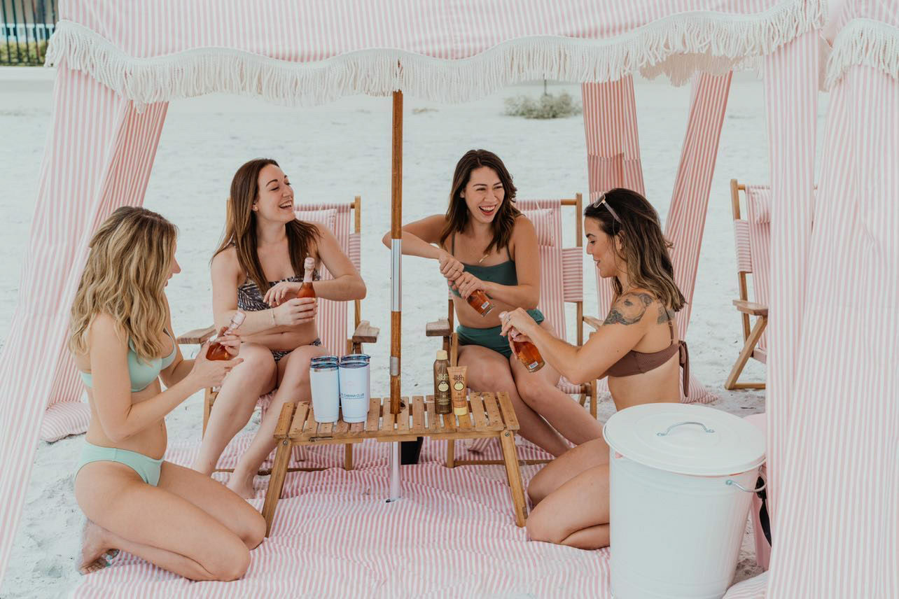 Four friends in bikinis laughing and opening rosé bottles inside a pink-and-white striped beach cabana on white sand, gathered around a small wooden table with tumblers, sunscreen, and a white cooler.