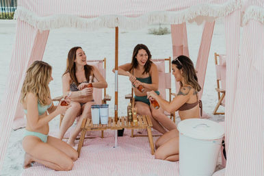 Four friends in bikinis laughing and opening rosé bottles inside a pink-and-white striped beach cabana on white sand, gathered around a small wooden table with tumblers, sunscreen, and a white cooler.