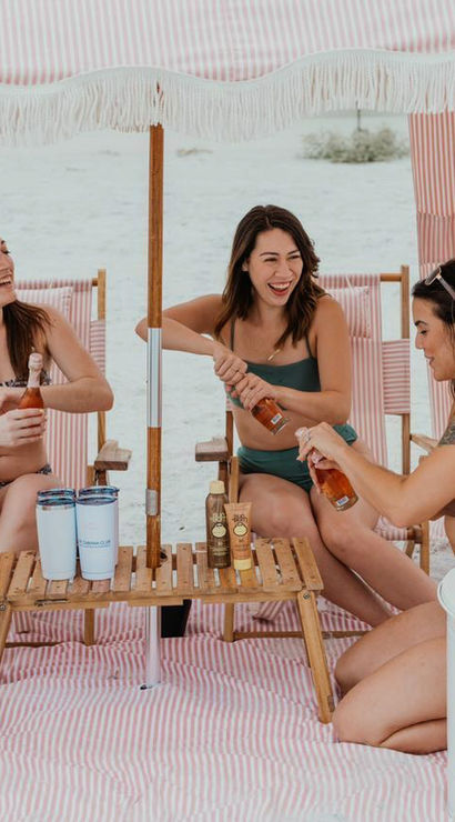 Four friends in bikinis laughing and opening rosé bottles inside a pink-and-white striped beach cabana on white sand, gathered around a small wooden table with tumblers, sunscreen, and a white cooler.