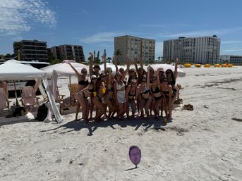 Group of women in swimsuits cheering under striped beach cabanas on a sunny sandy shore with oceanfront condos and yellow cabana umbrellas in the background; a novelty face cutout on a stake in the foreground.
