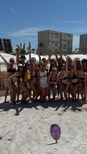 Group of women in swimsuits cheering under striped beach cabanas on a sunny sandy shore with oceanfront condos and yellow cabana umbrellas in the background; a novelty face cutout on a stake in the foreground.
