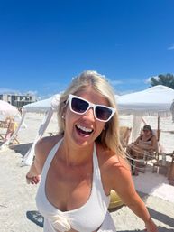 Smiling woman in white cat-eye sunglasses and textured white swimsuit on a sunny white-sand beach with cabana umbrellas and bright blue sky.