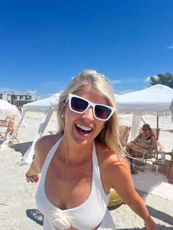 Smiling woman in white cat-eye sunglasses and textured white swimsuit on a sunny white-sand beach with cabana umbrellas and bright blue sky.