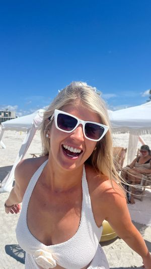 Smiling woman in white cat-eye sunglasses and textured white swimsuit on a sunny white-sand beach with cabana umbrellas and bright blue sky.