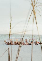 White-sand beach with a row of pale cabanas and umbrellas, lounge chairs and towels facing a calm turquoise ocean, framed by tall beach grasses in the foreground — relaxed seaside scene.