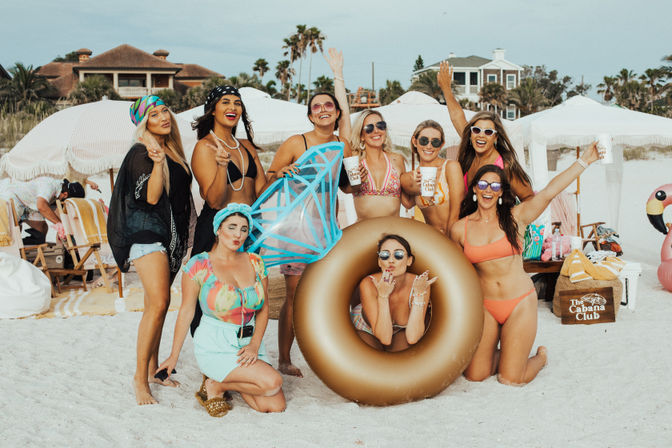 Group of smiling women in bikinis and beachwear posing at a sunny sand beach party with umbrellas, palm trees, coastal houses, a gold inflatable ring and novelty float — lively beachfront scene