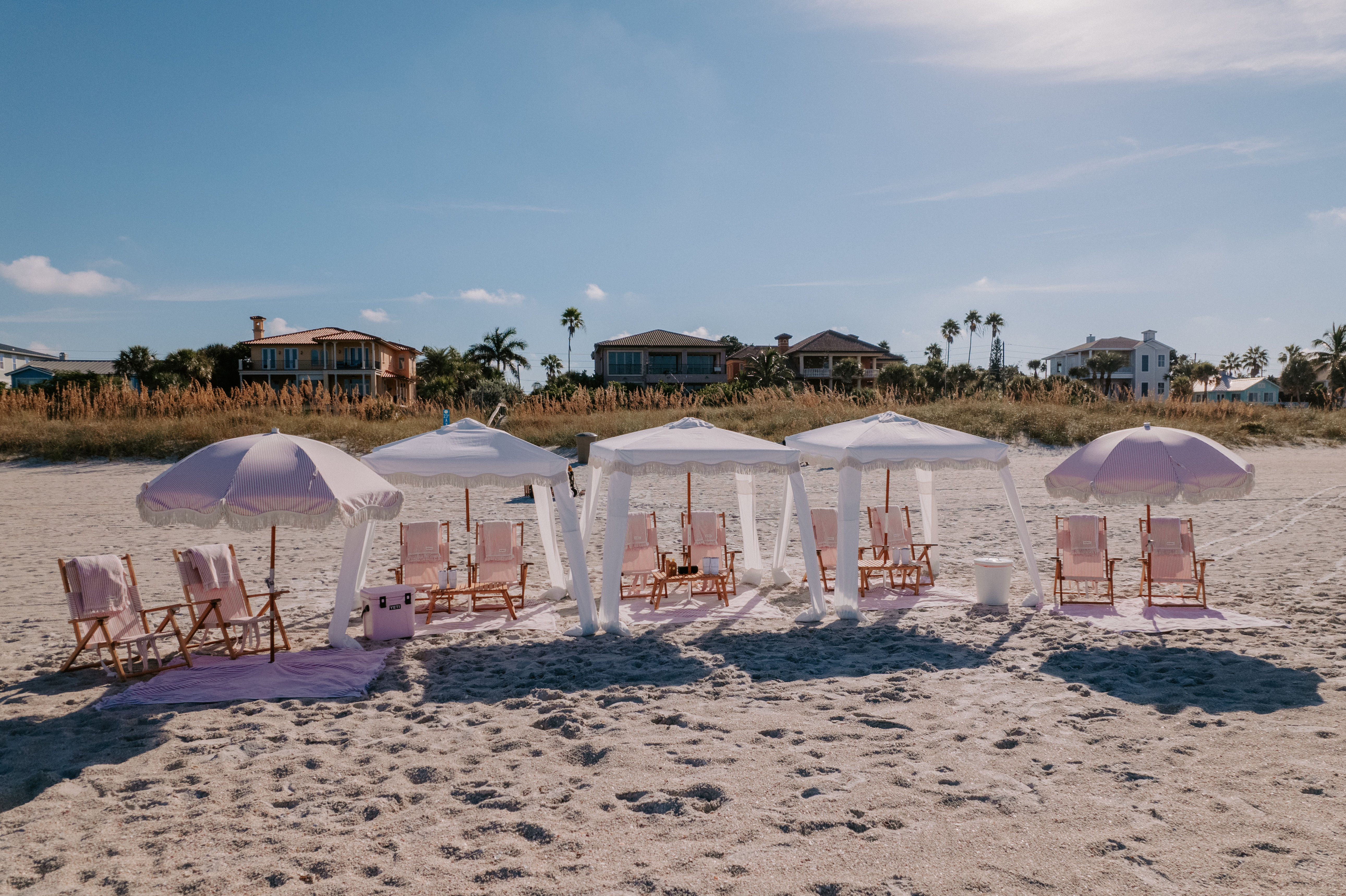 Sunlit sandy beach with pastel pink umbrellas and white canopies shading wooden lounge chairs, framed by dunes, palm trees and beachfront houses.