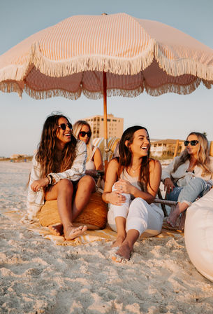 Four friends laughing under a pink striped fringed beach umbrella on a sandy beach at sunset, relaxed summer vibes with sunglasses and coastal buildings in the background.