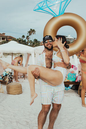 Sunlit beach party on a sandy coast with a man lifting a woman who kisses his cheek; both in pastel swimwear, oversized gold diamond-shaped inflatable ring and palm trees in the background, summer vacation vibe.