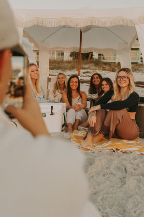 Group of friends sitting on a striped towel under a fringed beach canopy on a sandy shore at golden hour, smiling as someone snaps their photo beside a cooler.
