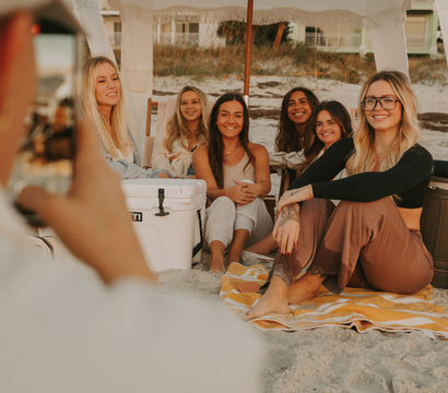Group of friends sitting on a striped towel under a fringed beach canopy on a sandy shore at golden hour, smiling as someone snaps their photo beside a cooler.