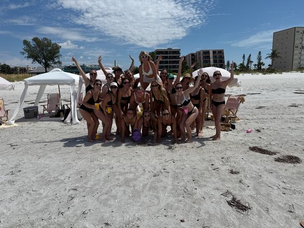 Large group of women in swimsuits posing and cheering on a sunny white‑sand beach with striped cabanas, folding chairs and oceanfront condominiums in the background.
