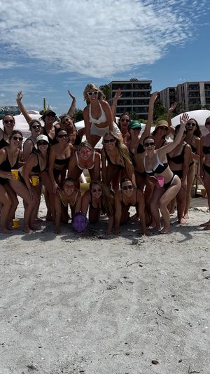 Large group of women in swimsuits posing and cheering on a sunny white‑sand beach with striped cabanas, folding chairs and oceanfront condominiums in the background.
