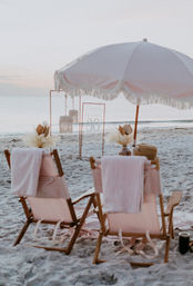 Soft pink-and-white striped beach chairs and a fringed umbrella on a sandy seaside at sunset, boho-style setup with pampas grass, hanging lanterns and a minimalist sign for a 30th celebration.