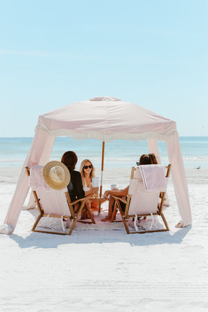 Three people relaxing under a pink-and-white striped beach canopy on white sand, seated in wooden chairs with drinks, calm turquoise ocean and clear blue sky beyond.