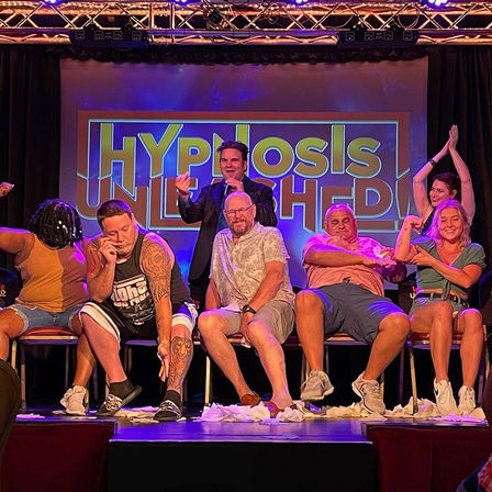 Energetic stage hypnotist entertains a row of adult volunteers on a lit comedy-club stage; participants laugh, wipe tears, flex and applaud with crumpled tissues scattered on the floor.
