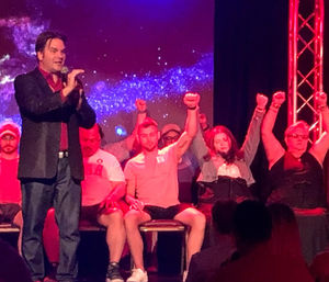 Performer on a theater stage with a microphone while seated volunteers raise their arms and appear hypnotized under red-purple stage lights, audience silhouettes in the foreground — live entertainment show.