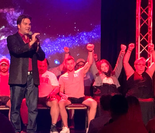 Performer on a theater stage with a microphone while seated volunteers raise their arms and appear hypnotized under red-purple stage lights, audience silhouettes in the foreground — live entertainment show.