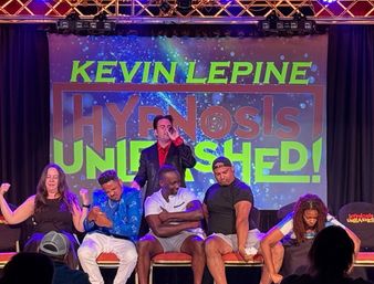 Live stage hypnosis show — performer with a microphone and five seated volunteers reacting dramatically against a colorful neon-lit backdrop.