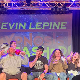 Five seated volunteers reacting dramatically on a lit theater stage during a live hypnosis show with colorful backdrop and stage lights.