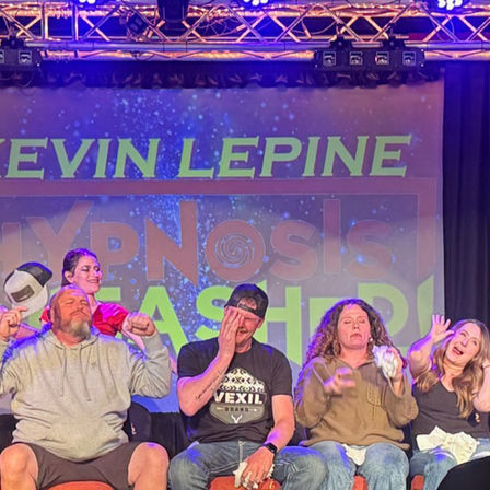 Five seated volunteers reacting dramatically on a lit theater stage during a live hypnosis show with colorful backdrop and stage lights.
