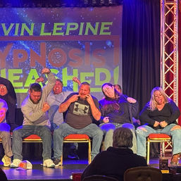 Row of seated volunteers making exaggerated faces and poses during a live stage hypnosis show with colorful "HYPNOSIS" backdrop and theatrical lighting.