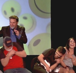 Emcee on an indoor stage with a large smiley-face projection behind him as three audience participants — a bearded man in a cap, a glasses-wearing man, and a laughing woman — double over laughing during a live comedy/entertainment show.