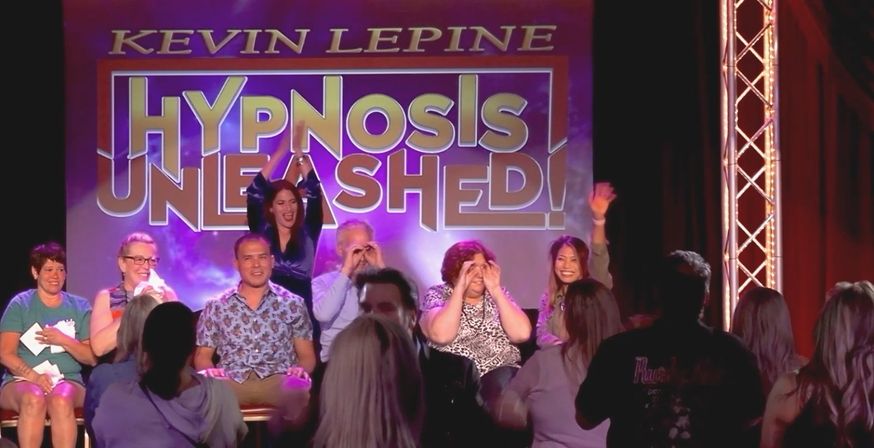 Live hypnosis show on a theater stage with seated volunteer participants under a large 'Hypnosis Unleashed' backdrop, purple stage lighting and an engaged audience watching.