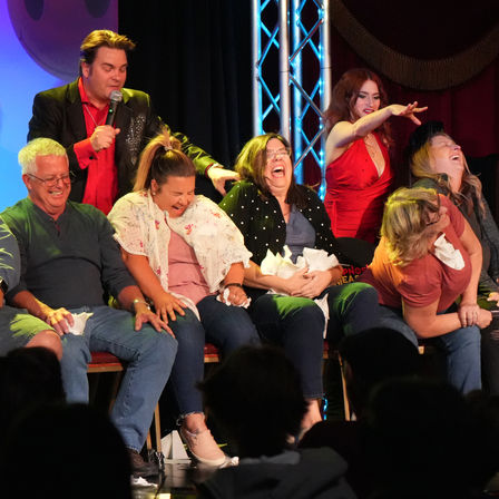 Live comedy hypnosis show on stage with seated volunteers laughing and clutching tissues while a microphone-wielding host and a red‑dressed assistant guide the act under stage lights.