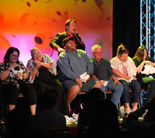 Row of laughing audience volunteers on a comedy club stage during a lively live entertainment show, performer with microphone engaging the crowd against a colorful backdrop.