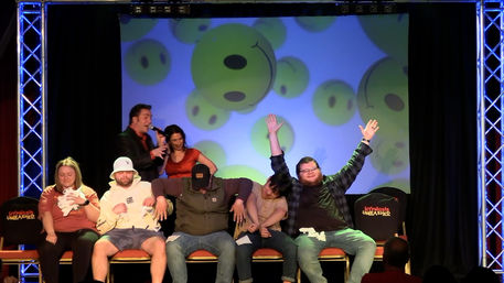 Live comedy hypnosis show on a small theater stage with volunteers seated in a row, two hosts with microphones, a backdrop of oversized smiley faces, and one participant cheering with arms raised.