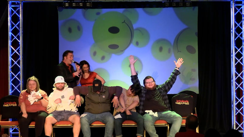 Live comedy hypnosis show on a small theater stage with volunteers seated in a row, two hosts with microphones, a backdrop of oversized smiley faces, and one participant cheering with arms raised.