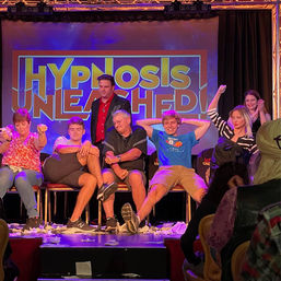 Live stage hypnosis show with audience volunteers seated across a theater stage, performer standing center in front of a bold “Hypnosis Unleashed” backdrop as participants slump, stretch and raise their arms in playful reactions.