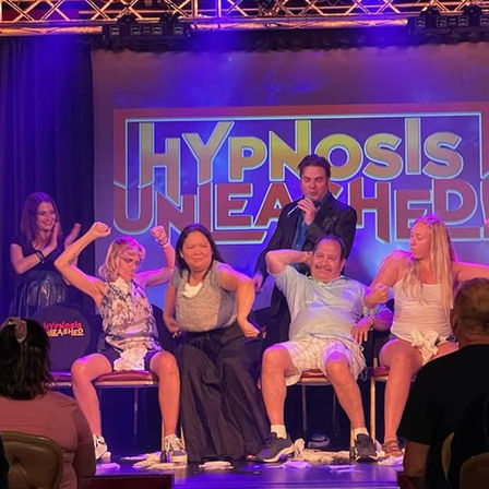 Live comedy hypnosis show on a colorful theater stage with a host holding a microphone and five seated volunteers striking energetic, hypnotized poses in front of a large "Hypnosis Unleashed" sign, colorful lights and audience visible.
