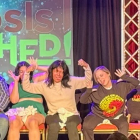 Three seated volunteers on stage making exaggerated faces and hand gestures under colorful lettered backdrop and stage lighting at a live comedy/improv show in an indoor theater